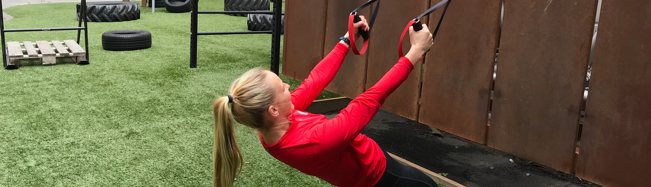 Tränare i röd Friskiströja gör ryggdrag i TRX i utegym.